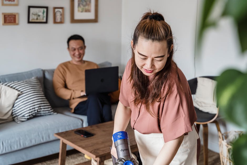 A woman smiling while vacuuming a living room, with a man working on a laptop in the background.